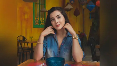 Smiling young woman in a denim shirt sitting at a cafe table with a warm, cozy atmosphere.