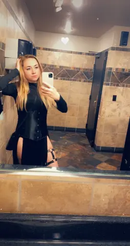 Blonde woman in a black long-sleeve top and corset outfit taking a mirror selfie in a tiled restroom.