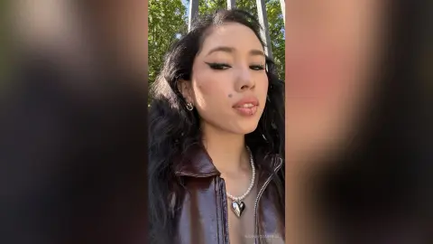 Young woman with winged eyeliner and pearl heart necklace wearing a brown leather jacket outdoors in sunlight.