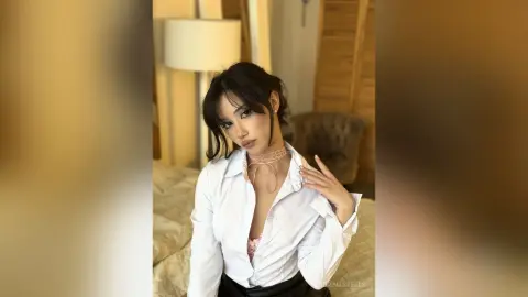 Young woman in a white button-down shirt and lace bra posing in a bright, sunlit bedroom.