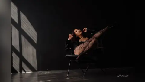 Elegant woman in black lace lingerie and leather jacket posing on a chair in a moody, dark studio setting.