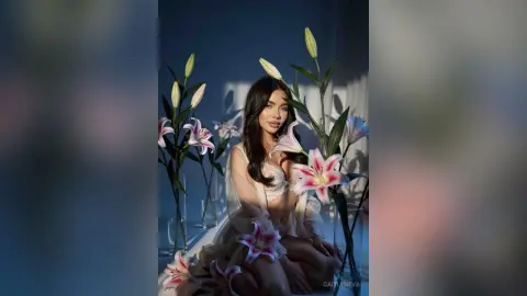 Beautiful woman in white lace lingerie sitting among pink lilies in a dreamy, soft-lit indoor setting.