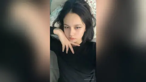 Young woman with dark hair in a black long-sleeve shirt relaxing on a bed with a moody, contemplative expression.
