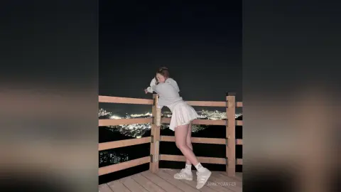 Young woman in a white sweatshirt and skirt posing on a balcony overlooking a glowing city skyline at night.