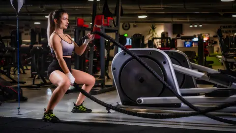 AmiraBelle wears a white sports bra and black shorts while doing battle rope exercises in a gym setting.