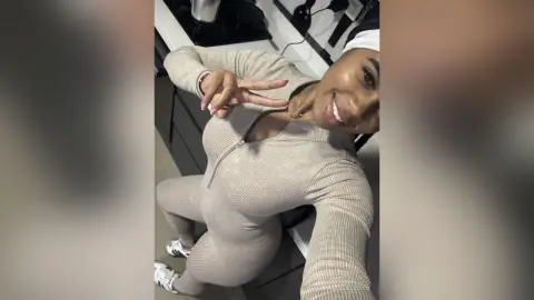 PamelaCreed smiles and flashes a peace sign while wearing a tight, long-sleeved tan ribbed jumpsuit or bodysuit.
