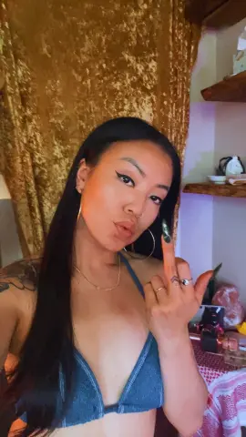 Serve_Cherry poses with long black hair and a denim bikini top, raising her middle finger against a gold velvet curtain background.