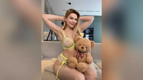 Smiling blonde woman in yellow lingerie sitting on a sofa with a brown teddy bear in a modern living room.
