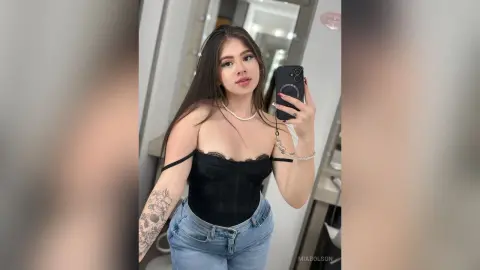 MiaBolson — Young woman in a black corset top and blue jeans posing for a mirror selfie in a bright room.