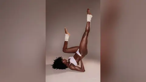Athletic Black woman in white bikini performing a graceful yoga stretch against a neutral studio background.