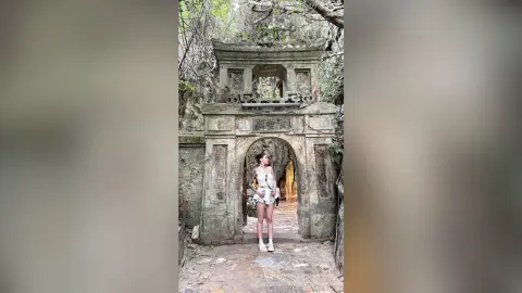 EmiliaRhodes poses in a short white dress standing in an ancient stone archway surrounded by trees.