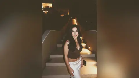 Young woman in a brown crop top and white skirt posing on outdoor stairs at night under warm lighting.