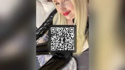 Smiling blonde woman in a black shiny outfit relaxing on a bed in a cozy indoor setting.