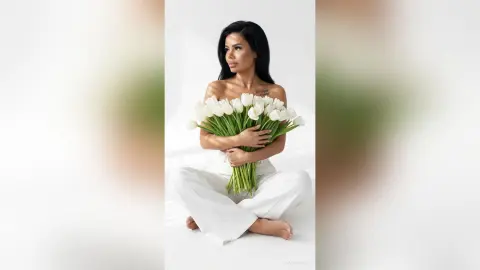 Elegant woman in white trousers holding a large bouquet of white tulips in a bright, minimalist setting.