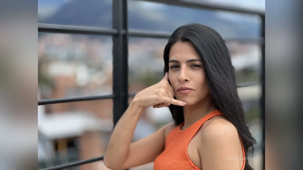RaquelWilde looks into the camera with long dark hair and an orange top against a blurred building background.