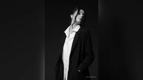 Elegant woman in a white shirt and black blazer posing in a moody, black and white studio portrait.