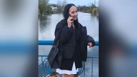 Smiling woman in a black puffer jacket talking on her phone by a peaceful lake at sunset.