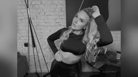 Blonde woman in a black crop top and leather pants posing in a modern loft studio setting.