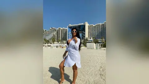 MissyJolie stands on a sandy beach wearing a short white button-down shirt and white sunglasses against a backdrop of large resort buildings.