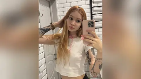 Blonde woman in a white t-shirt taking a mirror selfie in a modern bathroom with white subway tiles.