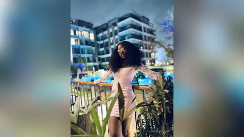 Stylish Black woman in a pink long-sleeve dress posing on a balcony overlooking modern city buildings at dusk.