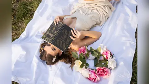 Dreamy woman in a beige linen dress reading a book on a white blanket in a sunny meadow with a rose crown.