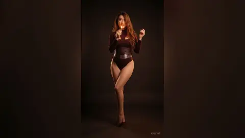Elegant woman in a dark burgundy bodysuit holding a lit match against a moody black studio background.