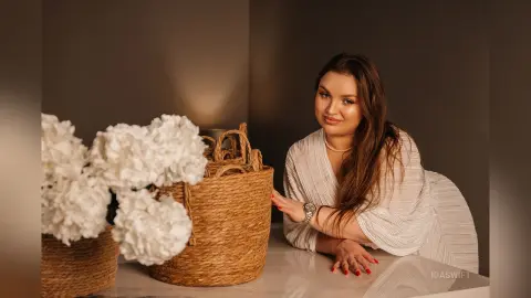 Smiling woman in a white pleated blouse sitting by white flowers and woven baskets in a warm, cozy setting.