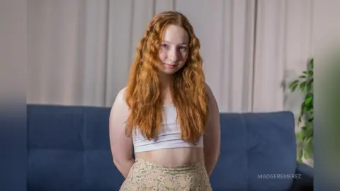 Smiling redhead woman in a white crop top and floral skirt sitting on a blue sofa in a bright room.