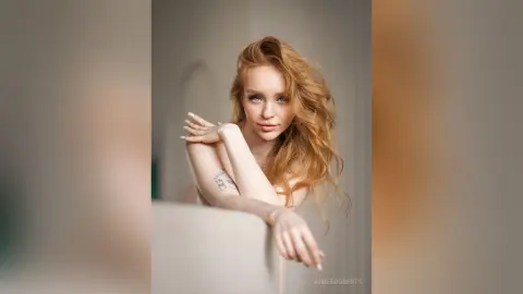 Blonde woman with long wavy hair posing gracefully in a soft, minimalist indoor setting with a serene mood.