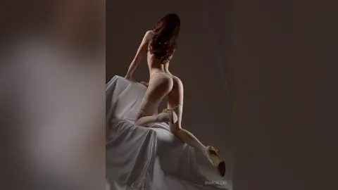 Elegant woman in lace lingerie and high heels posing gracefully on white draped fabric in a moody studio setting.