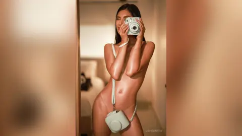 LucyMartin holds a white camera up to her face, posing naked in a bright room while showing her bare chest and stomach.
