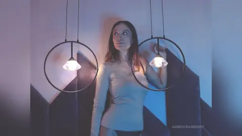 Young woman in a beige ribbed top posing between glowing circular pendant lights in a moody blue setting.