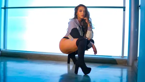 Fashionable woman in a grey shirt and black boots posing in a bright, modern blue-lit studio setting.