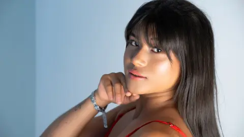 Portrait of a young woman with long dark hair and bangs wearing a red top in a bright studio setting.
