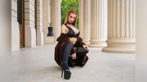 DeviNessa poses in black lace lingerie and a brown fur coat while kneeling on a stone floor between white columns.