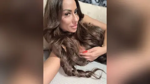 Smiling woman with long wavy brown hair relaxing on a white bed in a cozy indoor setting.