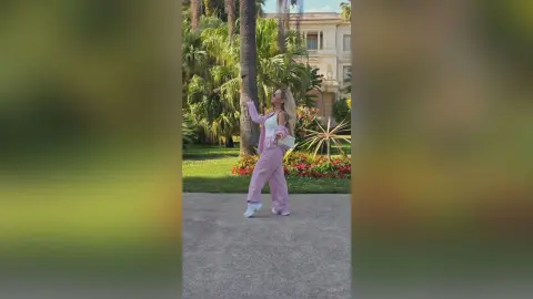 Cheerful woman in pink linen suit posing in a sunny tropical garden near a luxury villa.