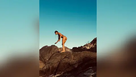 Fit woman in bikini standing on rocky mountain terrain under a clear blue sky in a serene outdoor setting.