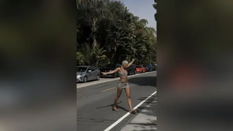 Cheerful woman in a striped bikini walking along a sunny tropical street lined with palm trees.