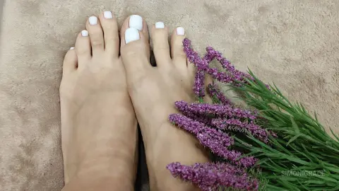 SimoniGrace shows off her white painted toenails resting on a tan fuzzy rug next to a bunch of purple flowers.