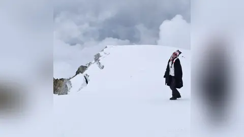 Adventurous traveler in black winter gear standing on a vast snowy mountain peak under a cloudy sky.