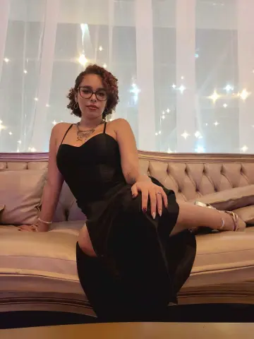 dominamoon — Elegant woman in a black cocktail dress sitting on a beige sofa in a dreamy, glowing room.