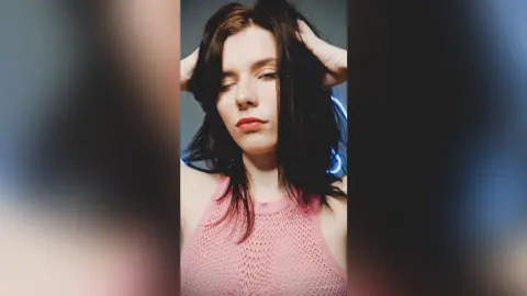 Young woman with dark hair in a pink mesh tank top posing with eyes closed in moody studio lighting.