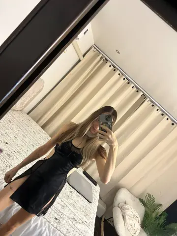 Young woman in a black satin slip dress taking a mirror selfie in a bright, modern bedroom.