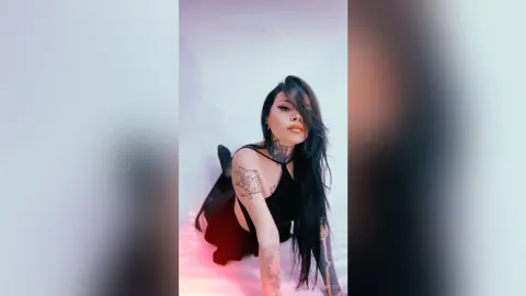 KatherinBesse poses with long black hair, wearing a black tank top and showing off her tattoos against a plain white background.
