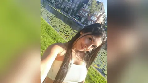 Young woman in a white tank top smiling by a scenic canal in a European city on a sunny day.