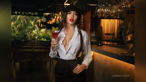 Glamorous woman in a white shirt and black suspenders holding a cocktail in a moody, dimly lit lounge bar.