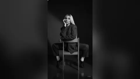 Elegant woman in a black power suit sitting on a chair in a moody, minimalist black and white studio setting.