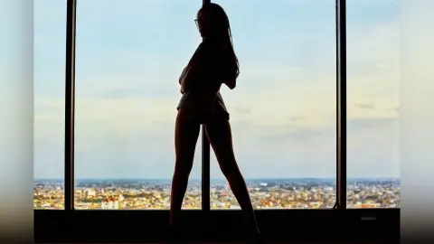 JessieLiu stands in silhouette against a large window overlooking a city skyline, showing her body shape and long hair.